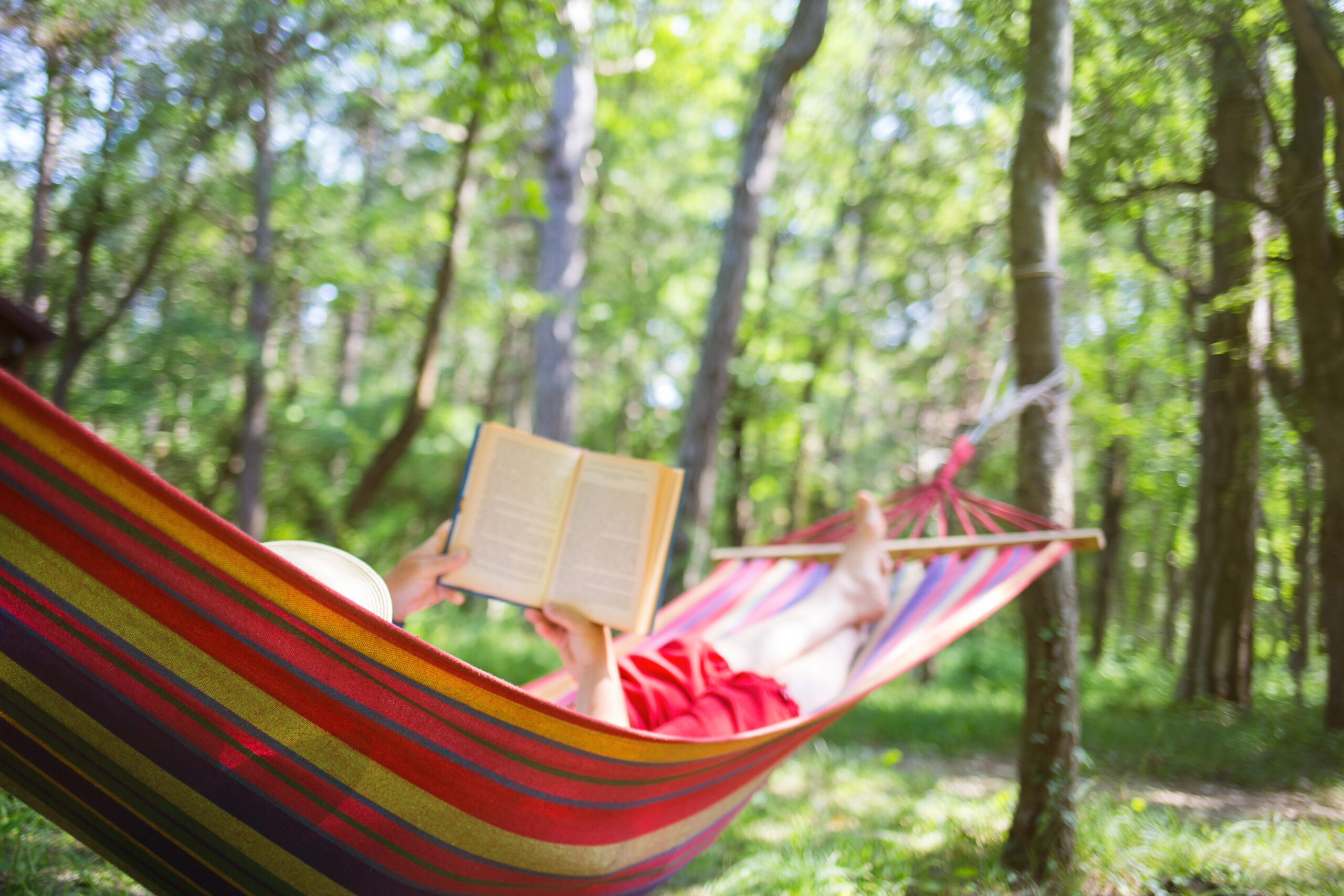 Photo of a white woman reading a book in a red and yellow striped hammock, surrounded by trees.