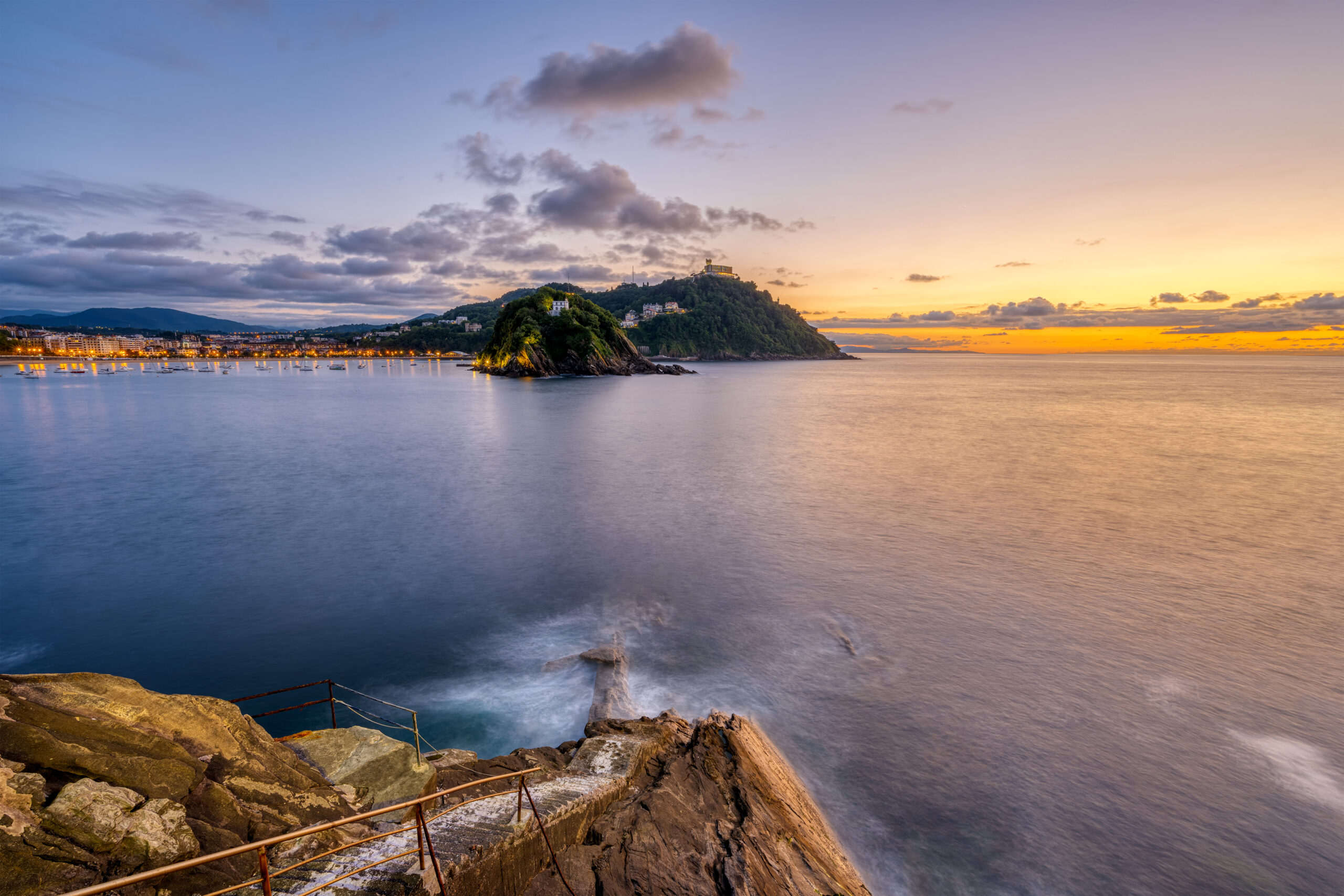 Photo of the bay in San Sebastian at sunset, with hills and the lights of the harbor in the background.