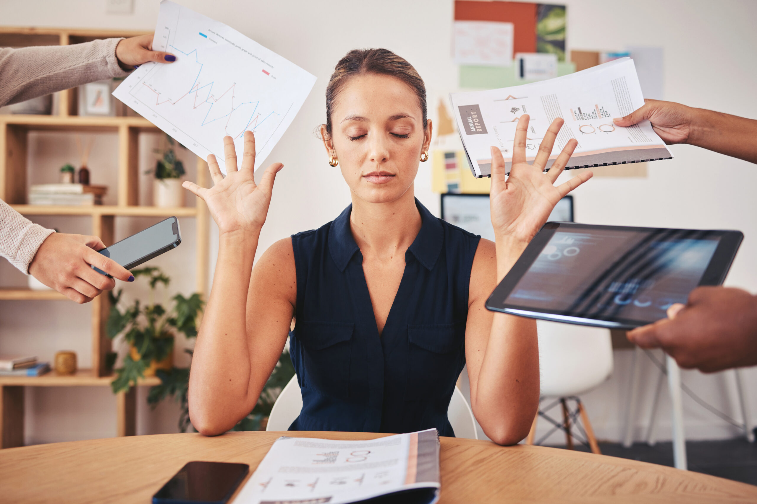 Photo of a white woman with her eyes closed and her hands up in a "stop it" gesture, as several hands come in from outside the frame holding spreadsheets, a tablet and a phone.