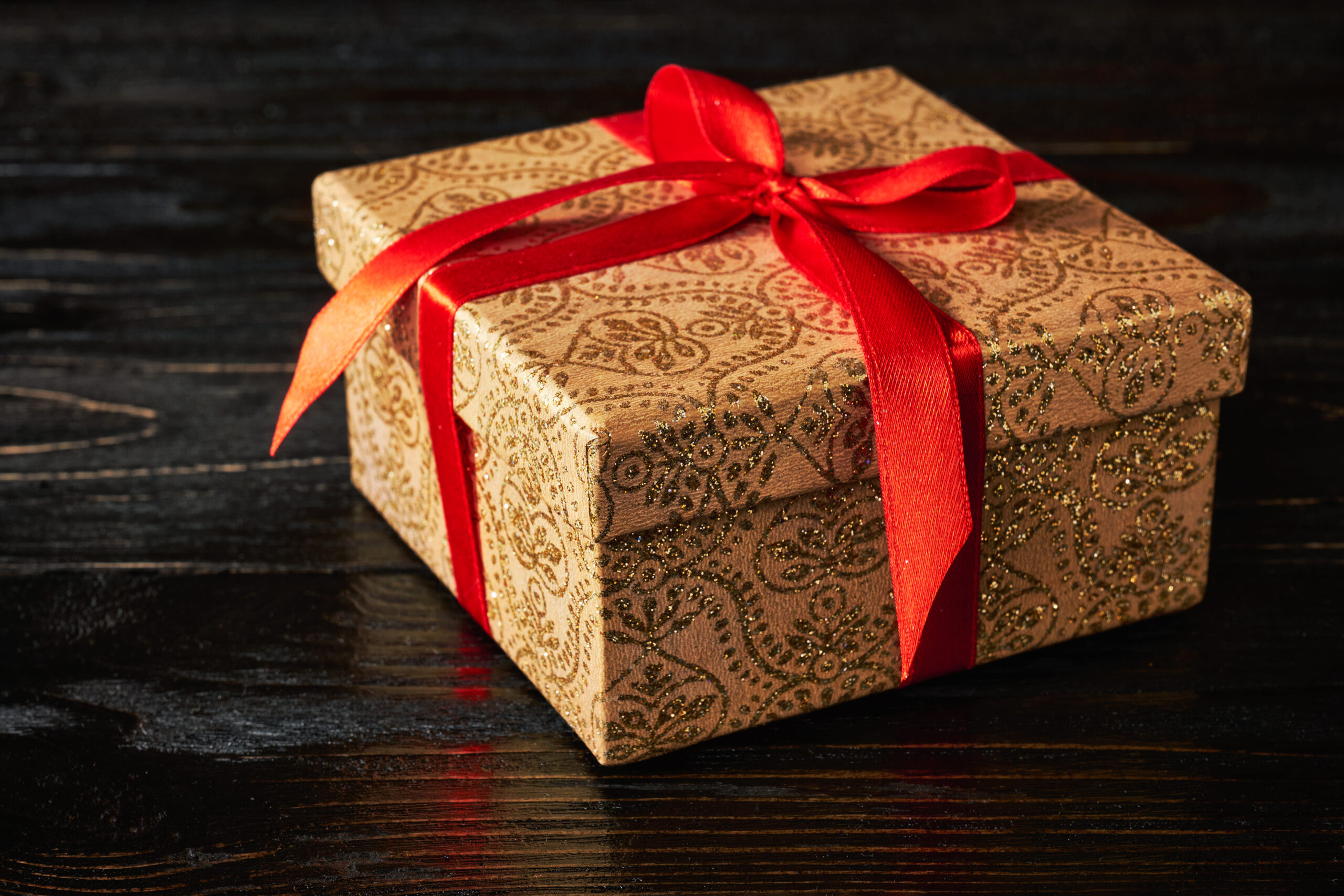 Photo of a gold gift box with a red bow, sitting on top of a dark wooden table.