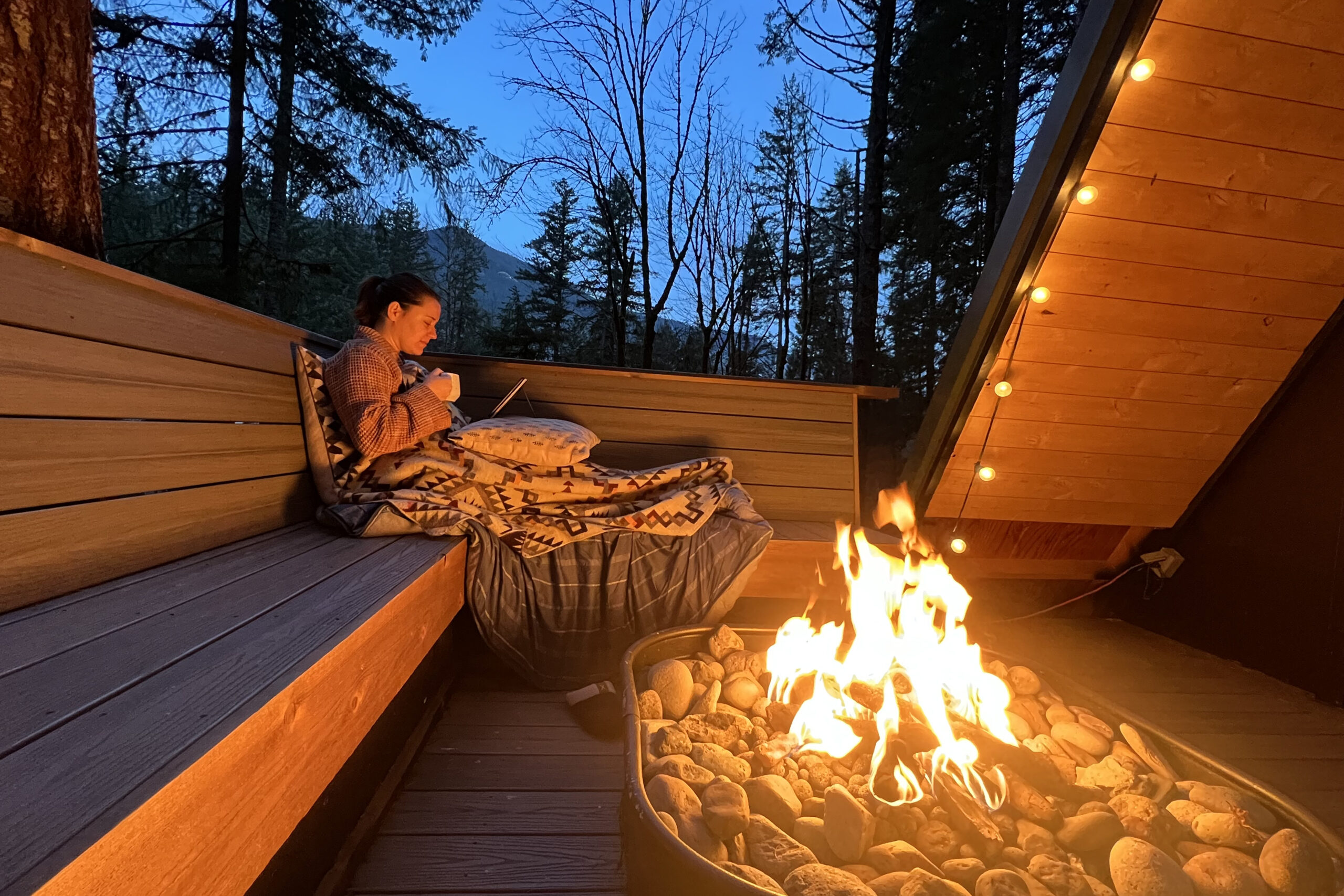 Photo of Maegan sitting on an outside bench, covered in blankets, reading a book next to the fire.