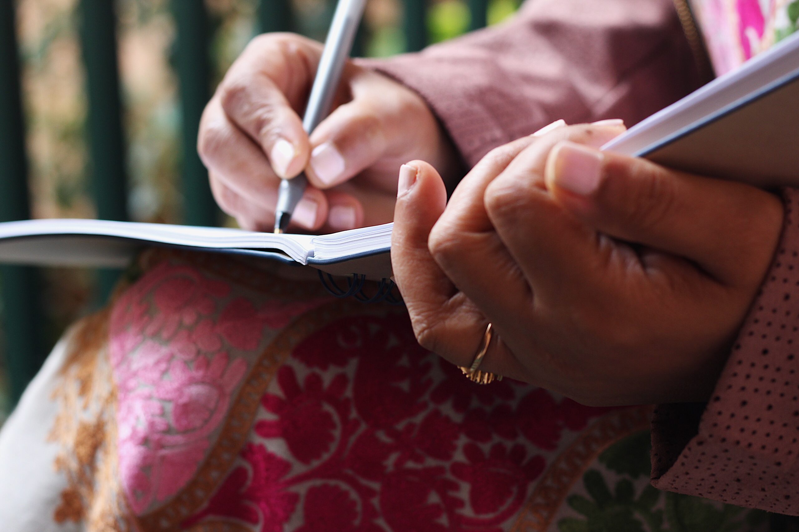 Closeup photo of a woman's hands holding a journal and pen, writing while wearing a pink shirt and patterned skirt.