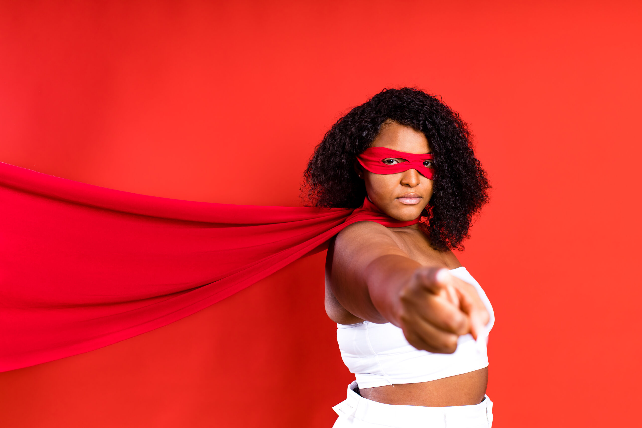 Photo of a Black woman with dark, curly hair, wearing a white top and pants and a red cape and superhero eye mask in front of a solid red background.