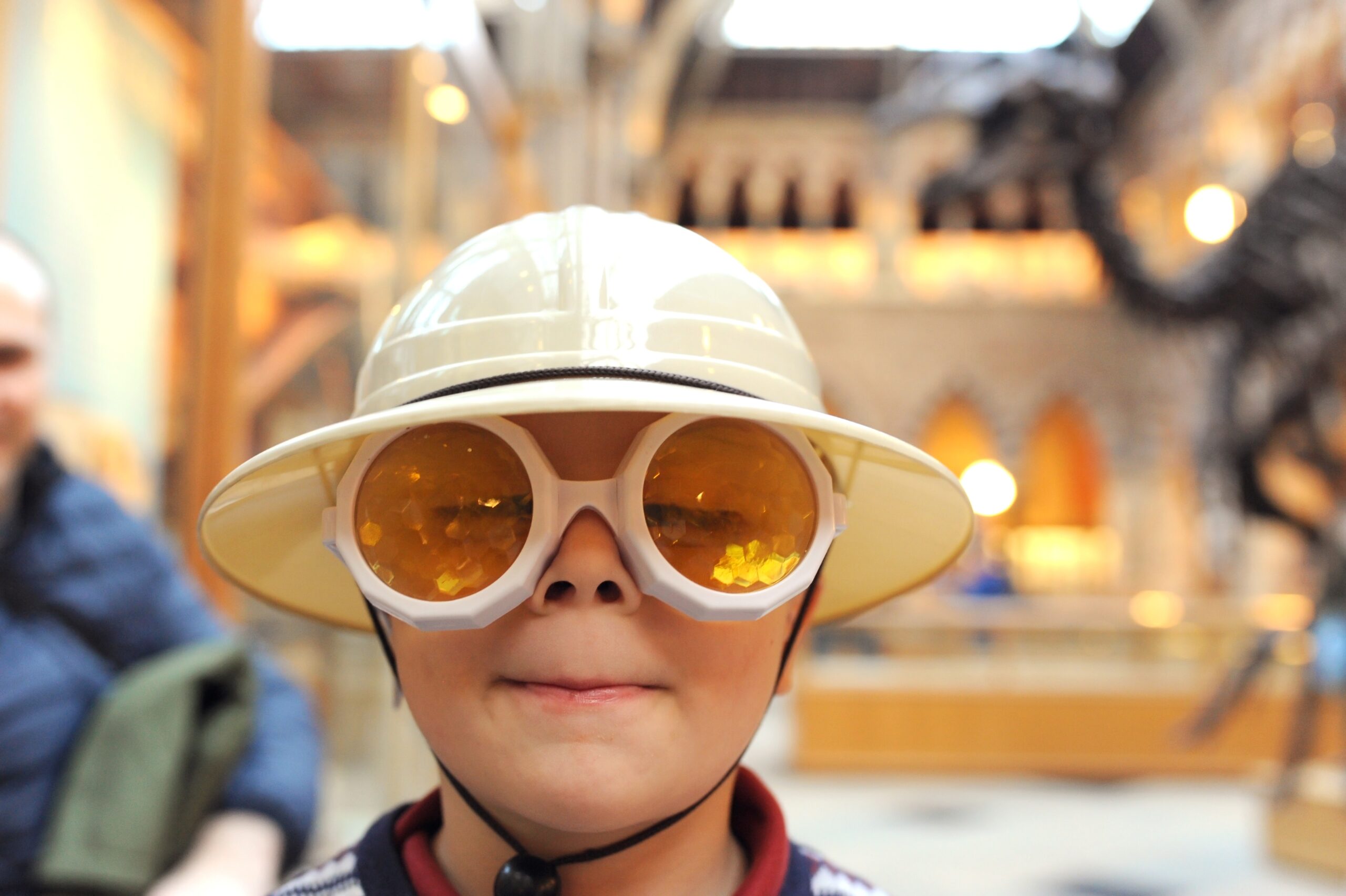 Photo of a young person wearing a safari hat and yellow goggles, with an exhibit of fossils in the background behind them.