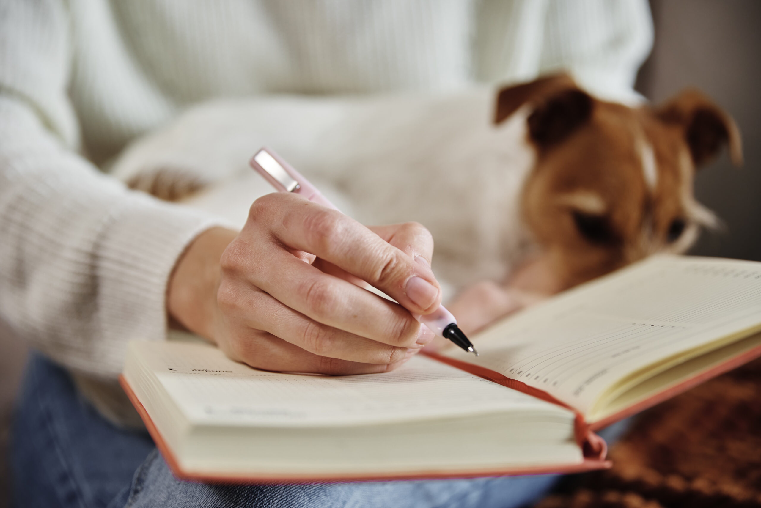 Photo of a person with a white shirt and light skin writing in a journal with a dog on their lap.