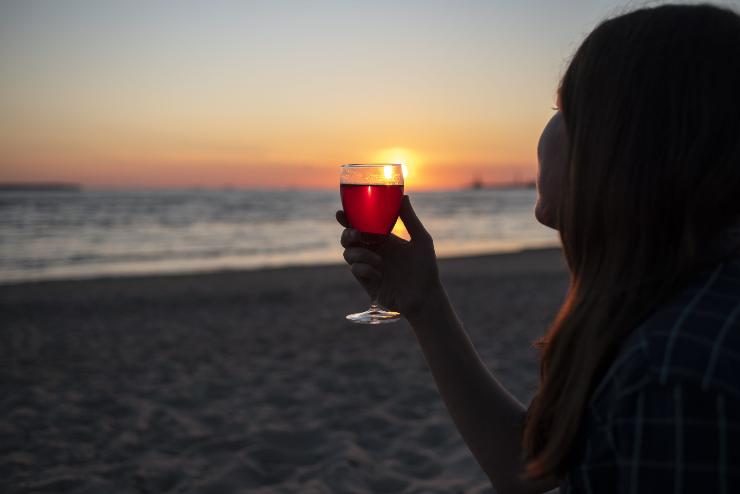 Photo of a woman's silhouette at dusk on the beach, holding a red wine glass just in front of the sun, making the red wine bright and lit up.