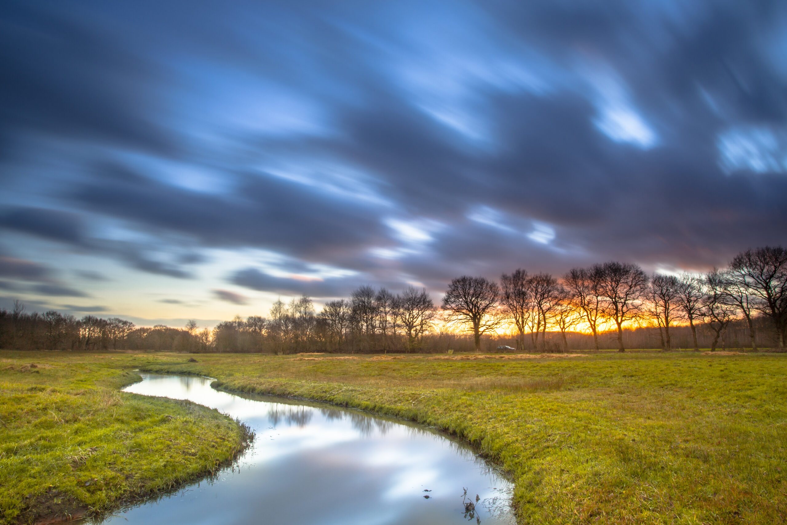 Photo of a stream running through a green, grassy meadow, with a line of trees and a sunset in the background, with the clouds in the sky blurry with motion.