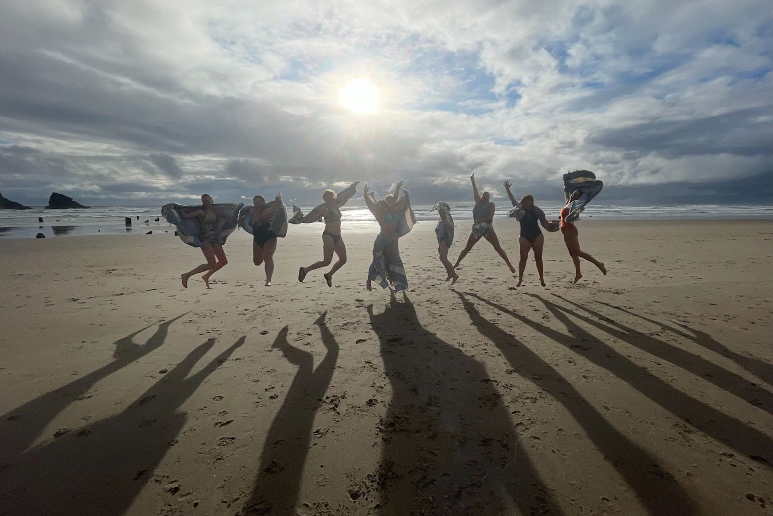 Photo of eight women silhouetted against the setting sun on the beach, all jumping with glee as their shadows stretch out toward the camera.
