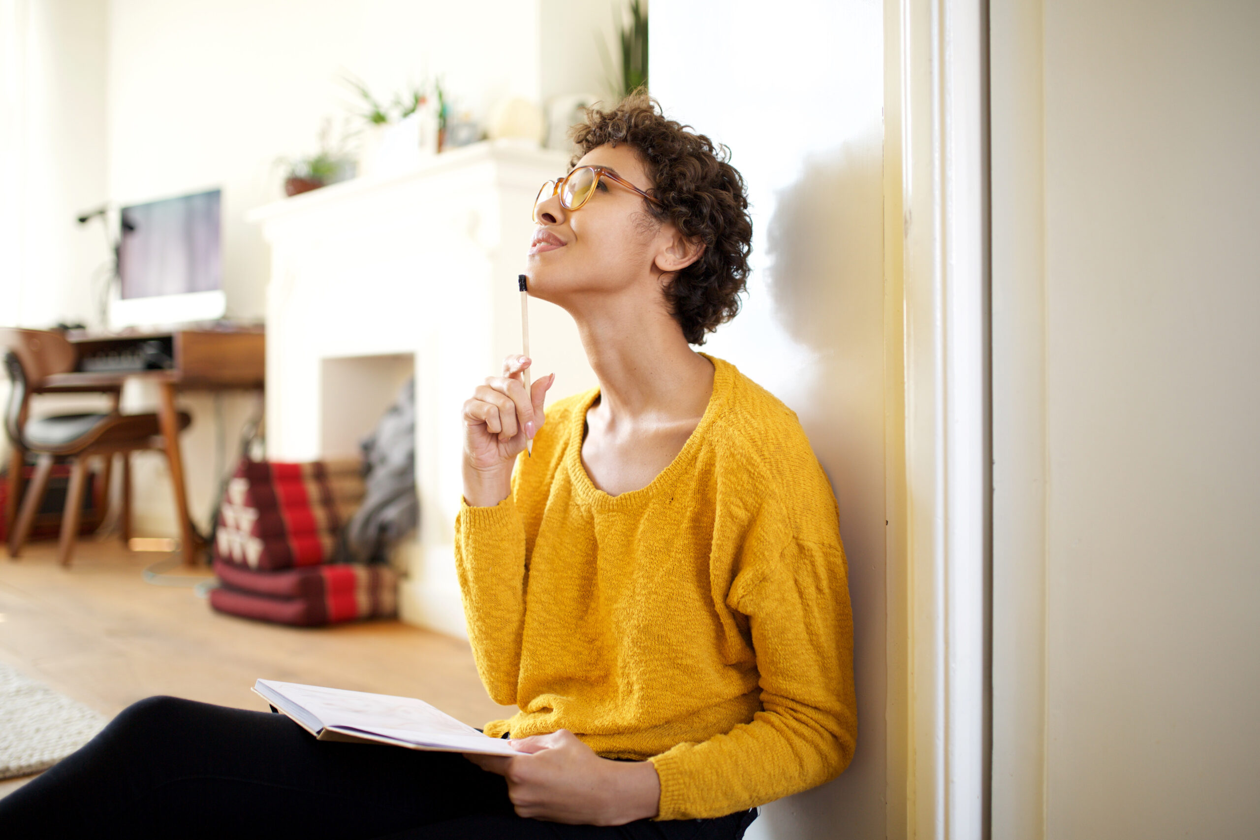 Photo of a woman with light brown skin, short, curly brown hair and glasses, wearing a yellow sweater and sitting in an inside doorway with a journal, holding a pen up to her chin in a pensive yet joyful pose.