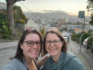 Image of Maegan, a white woman with shoulder length brown hair and glasses, and Linzy Bonham, a white woman with blonde hair and glasses, at the top of a steep hill overlooking the city of San Francisco at dusk.