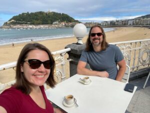 Photo of Maegan, a white woman with shoulder length brown hair and sunglasses, and her husband Jonathan, a white man with long brown hair and sunglasses, sitting in an outdoor cafe on the beach in San Sebastian, Spain.
