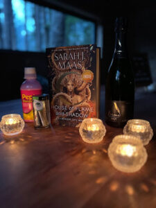 Photo shows Sarah J Maas' book, House of Flame and Shadow, standing upright on a table surrounded by candles, a jar of joints, a bottle of wine and a bottle of Pepto.