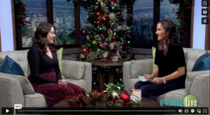 Image is a shot from Maaegan's appearance on a local tv talk show and features Maegan, a white woman with long brown hair in a burgundy dress, sitting on a couch opposite her host, another white woman with long brown hair in a black top and pants.