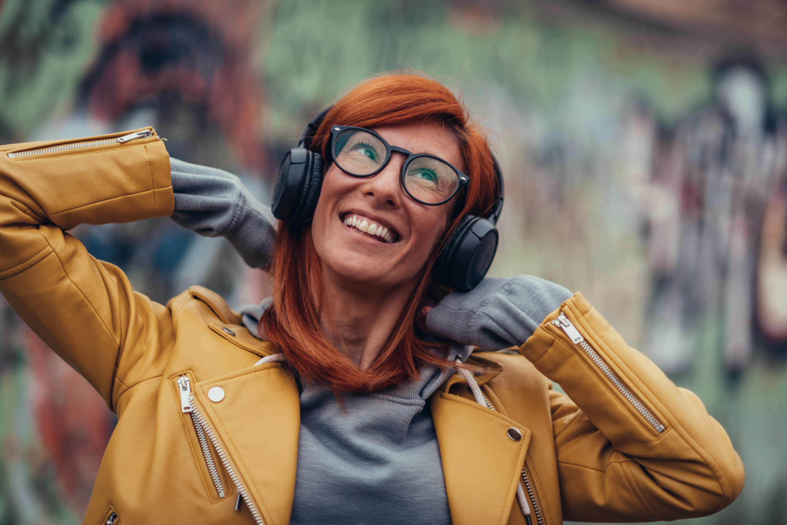 Photo of a woman with red hair and a yellow jacket, wearing headphones and smiling while covering her ears.
