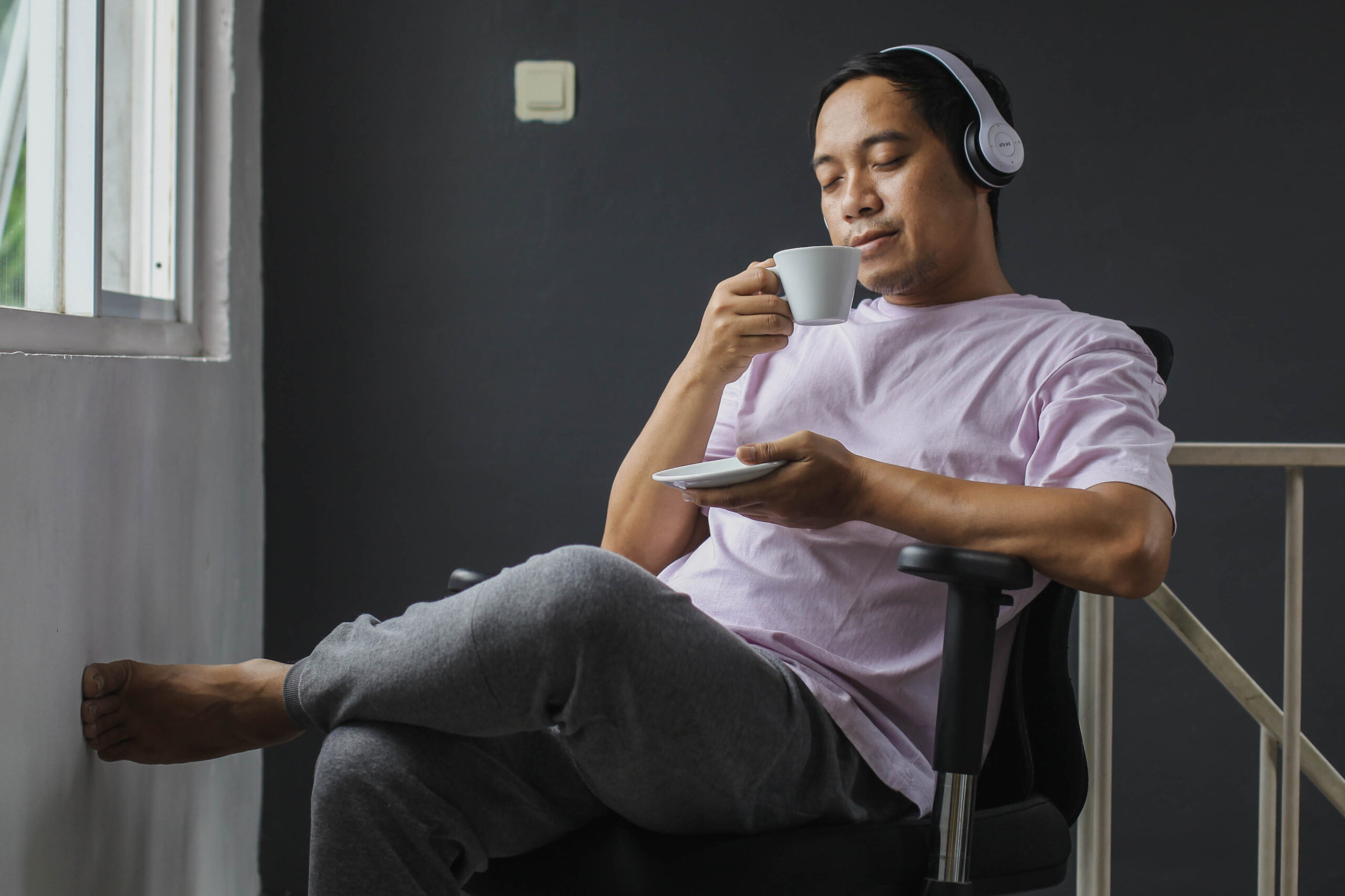 Photo of a Black man sitting in a chair, drinking coffee with headphones on and phone in hand.