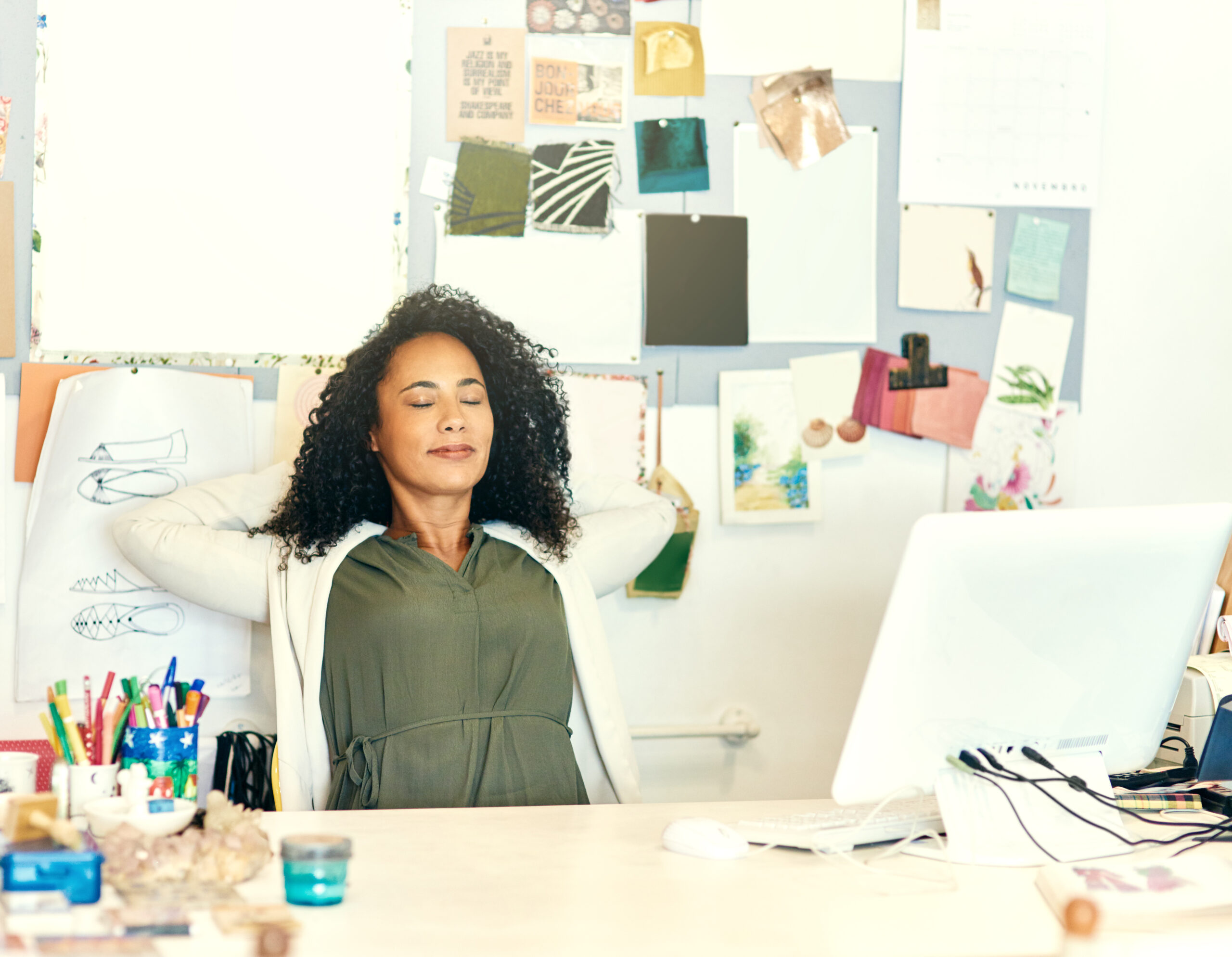 Photo of a Black woman, with long curly black hair, sitting back behind her desk, with her hands behind her head and her eyes closed.