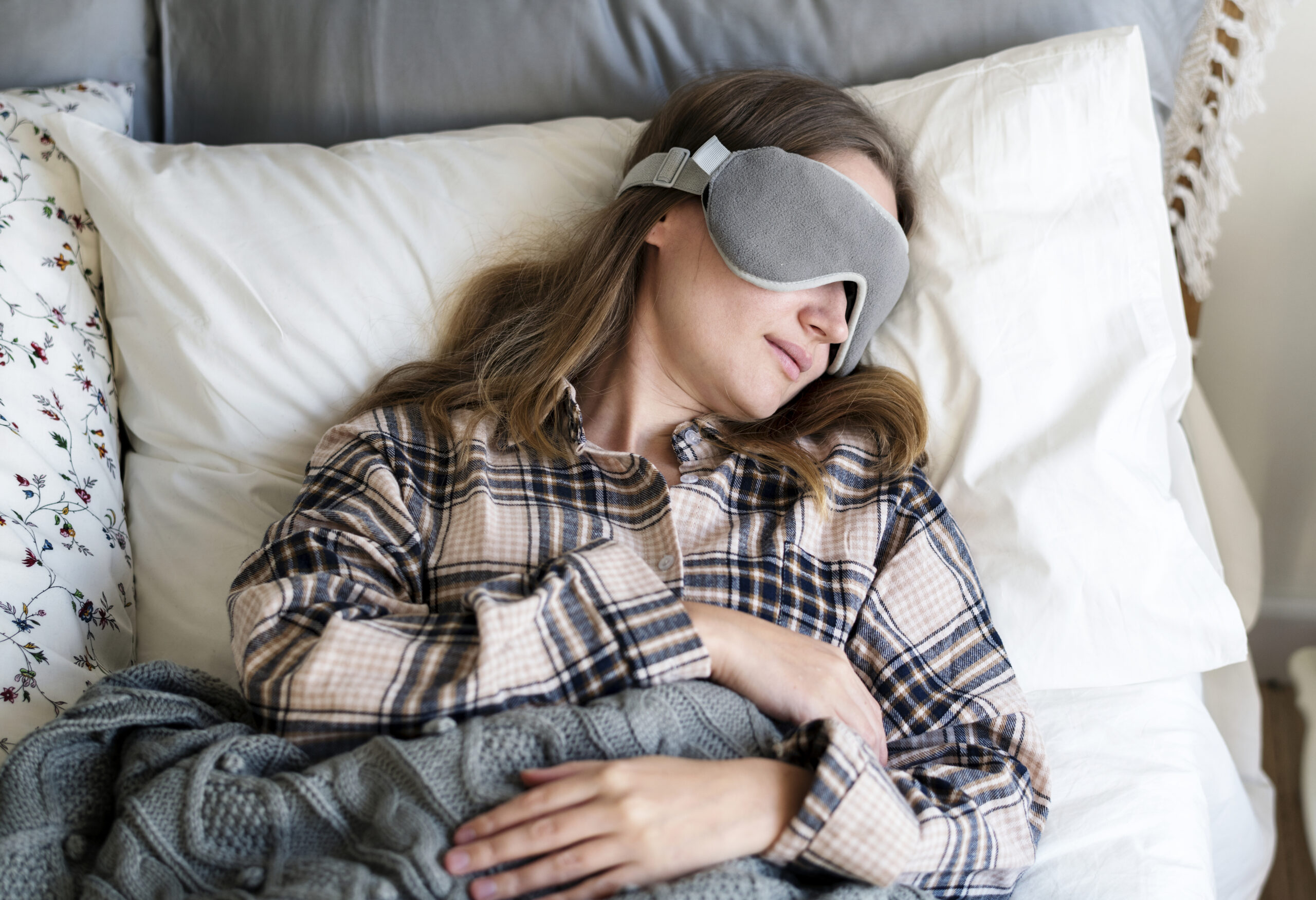 Photo of a white woman lying in bed in her pajamas, with an eye mask on and a blanket on top of her.