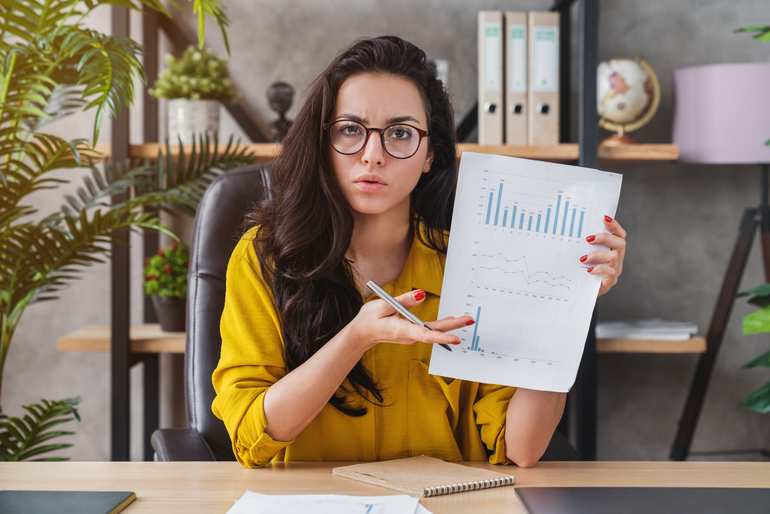 Photo of a Latina woman with long dark hair and glasses, sitting behind a desk and holding up a paper with graphs and charts on it.