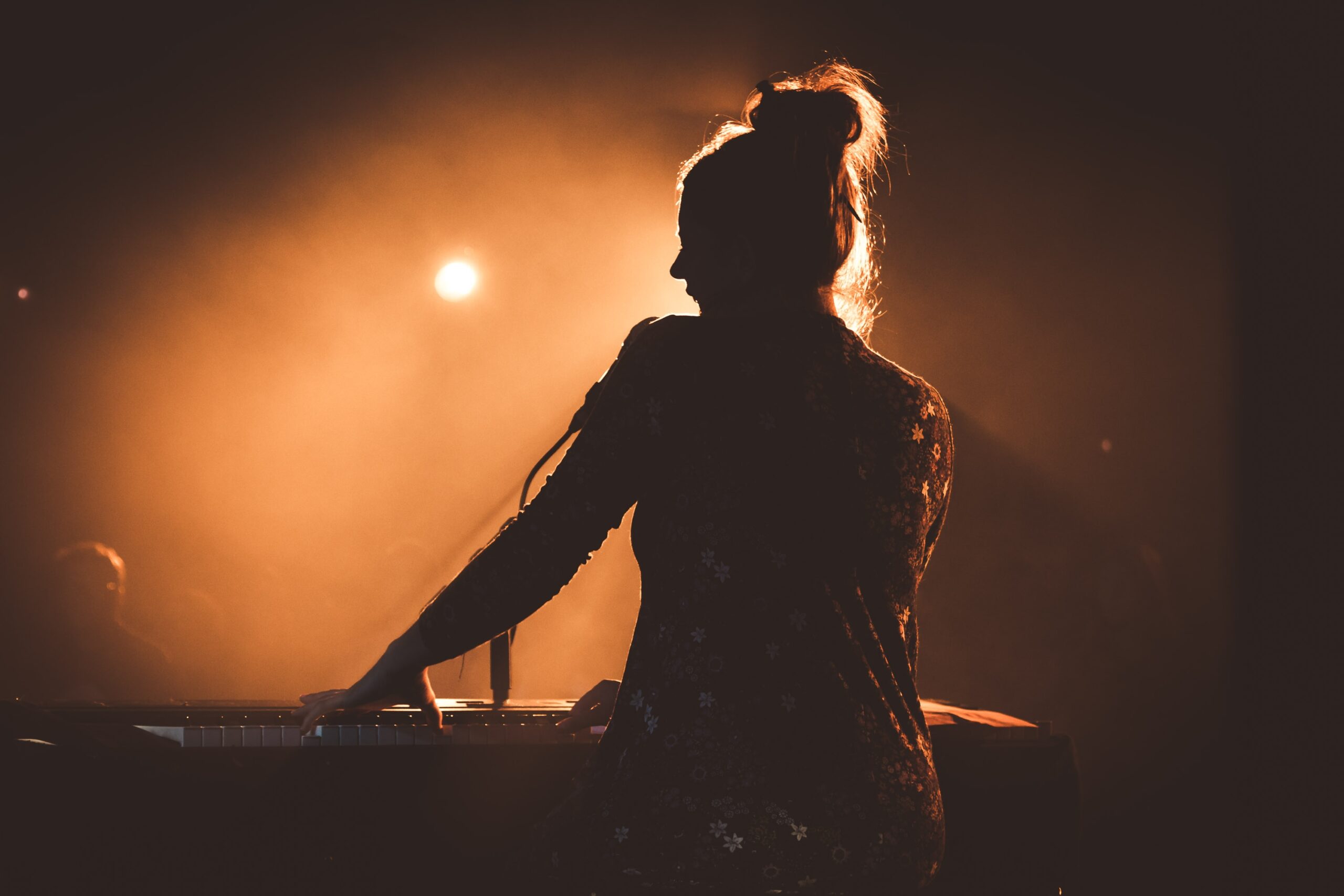 Photo of a silhouette of a woman playing piano and singing on stage with bright lights shining in the background.
