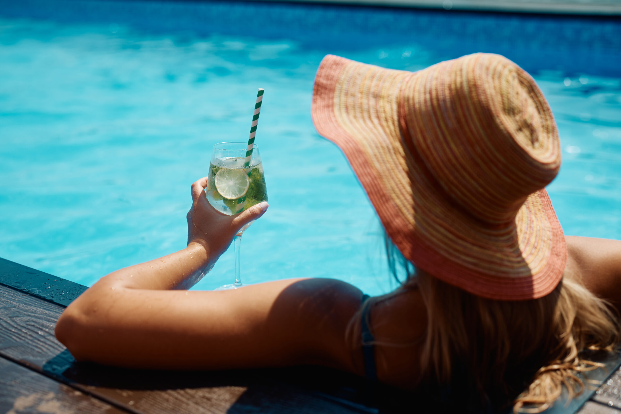 Photo of the back of a woman in a pool, wearing a sun hat with a margarita in her hand.