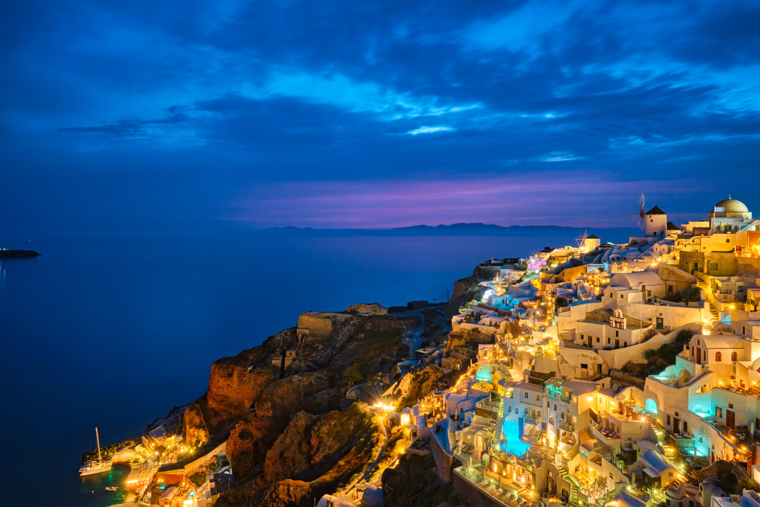 Photo of Santorini at night, with the houses lit up on the hill and the Mediterranean Sea in the background.