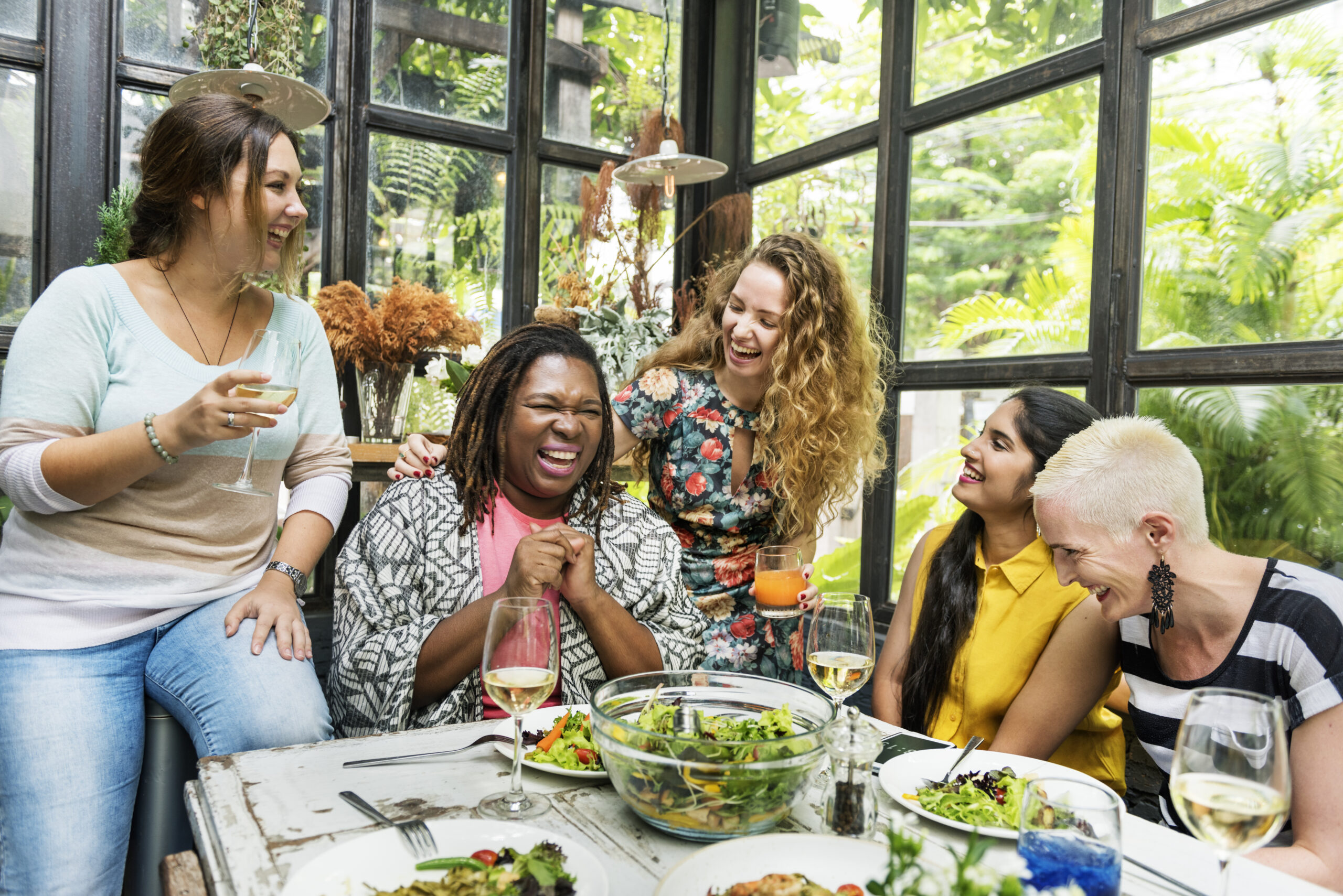 Photo of a diverse group of women laughing and sitting around a table with salads and glasses of wine.