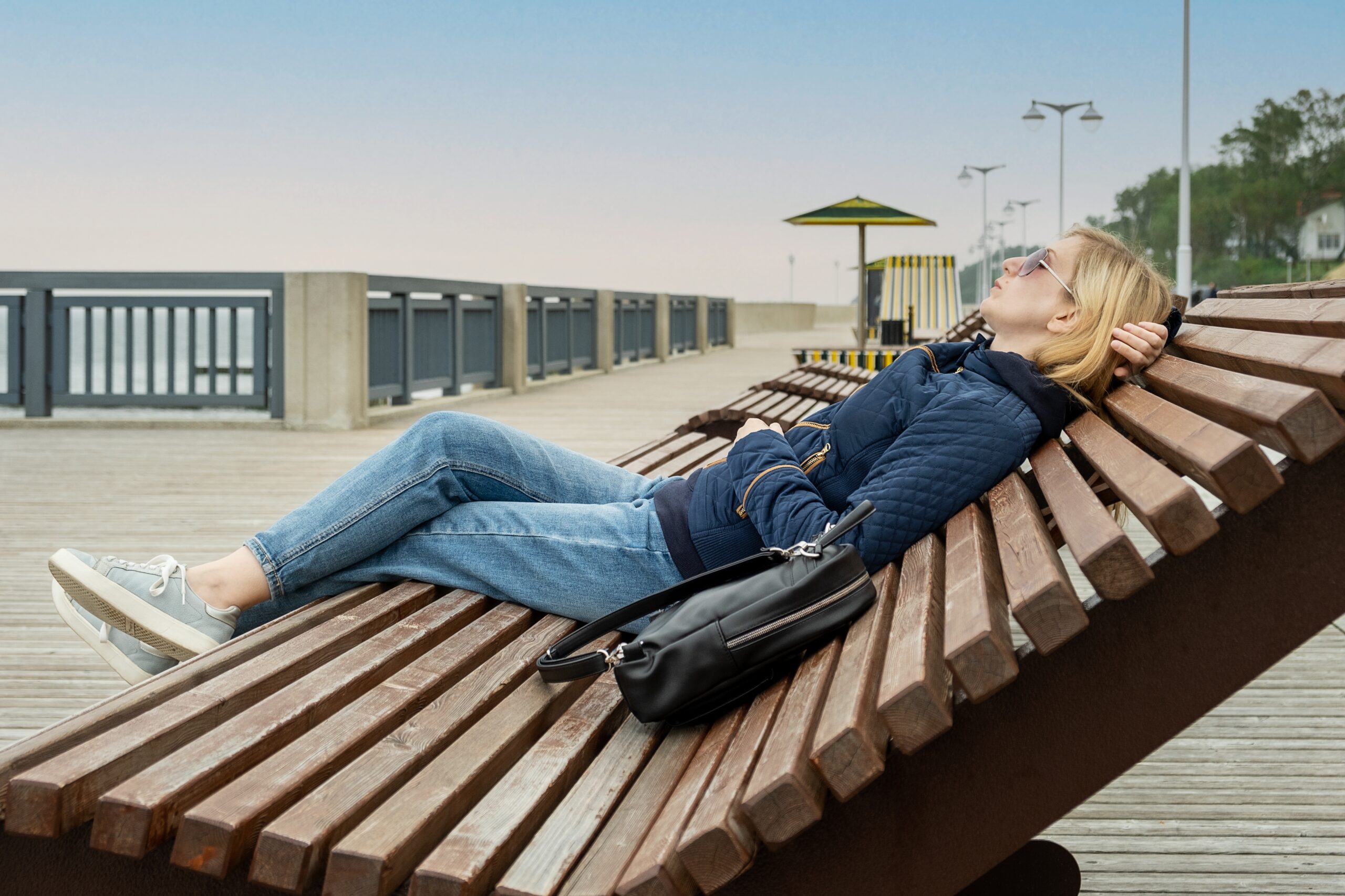 Photo of a white woman with blonde hair laid back on a lounge chair on a boardwalk with blue sky and the ocean in the background.
