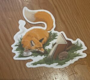 Photo of a sticker featuring a red fox wearing glasses and reading a book.