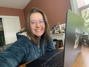 Image of Maegan, a white woman with medium length hair, sitting in front of her laptop dressed in a teal bathrobe, smiling at the camera.