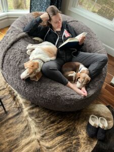Photo of Maegan, a white woman with brown hair, wearing sweats and reading a book in her LoveSak bean bag with 2 beagels snuggled next to her.
