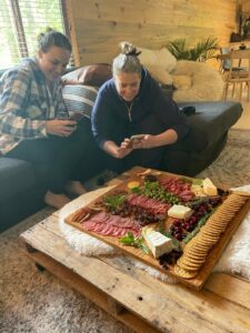 Photo of two women taking photos of a massive charcuterie board, covered in meats, cheeses, grapes, olives, cherry tomatoes, and crackers.