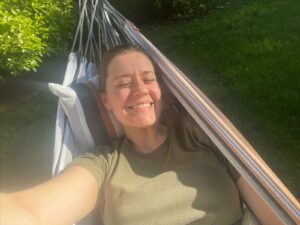 Maegan is lying in a hammock outdoors, smiling broadly with her eyes closed, enjoying the sunlight. She has brown hair pulled back and is wearing an olive green t-shirt. The hammock has a striped pillow behind her head. The background features green grass and bushes, indicating a garden or backyard setting. The sun is shining brightly, casting shadows and highlighting Maegan's content expression.