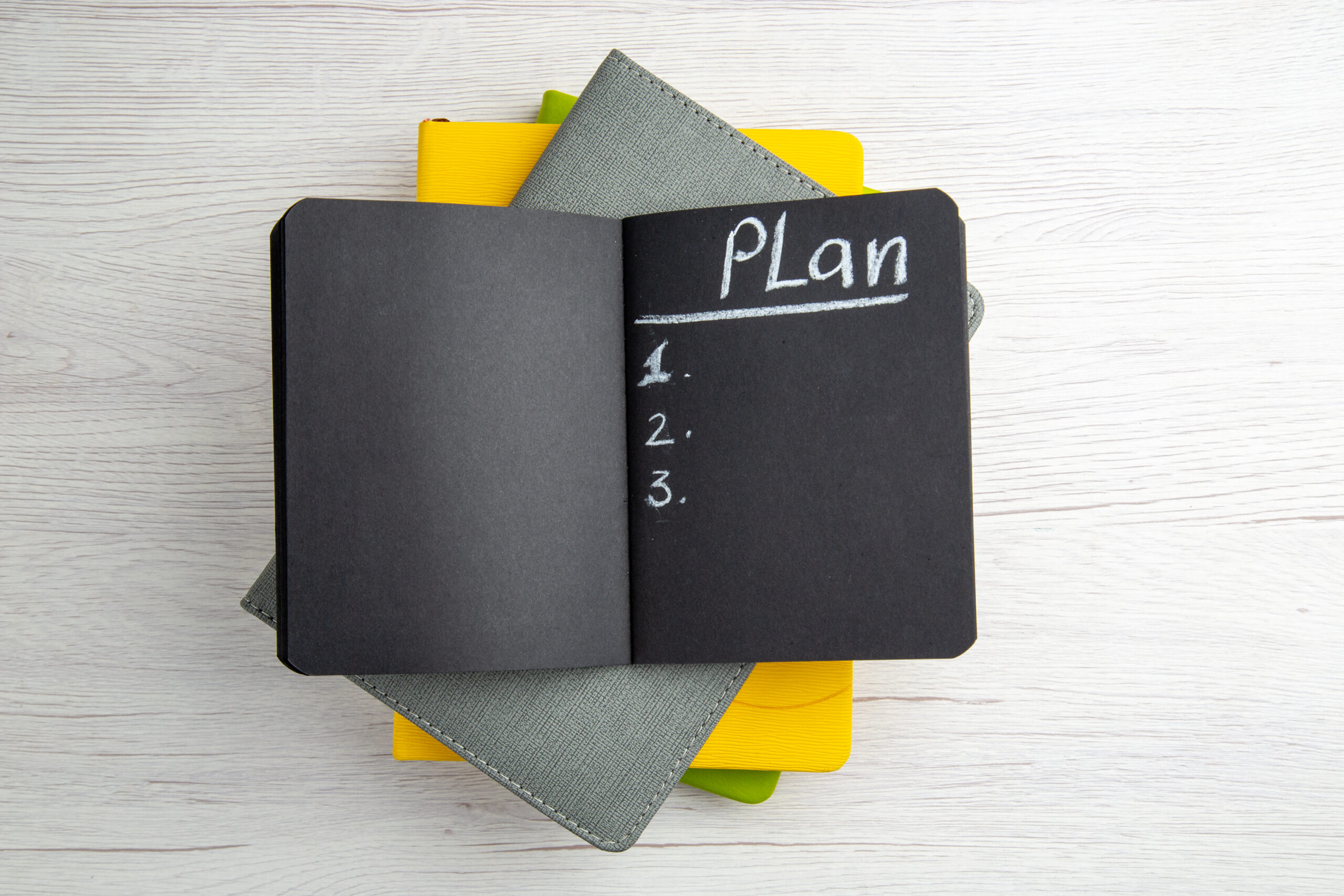 An open black notebook lies on top of a stack of closed notebooks with grey, green, and yellow covers, arranged on a light wood surface. The black notebook has the word "Plan" written at the top of the right-hand page in white chalk or chalk-like writing, followed by a numbered list with three items, all currently blank. The overall scene suggests planning or organizing tasks.