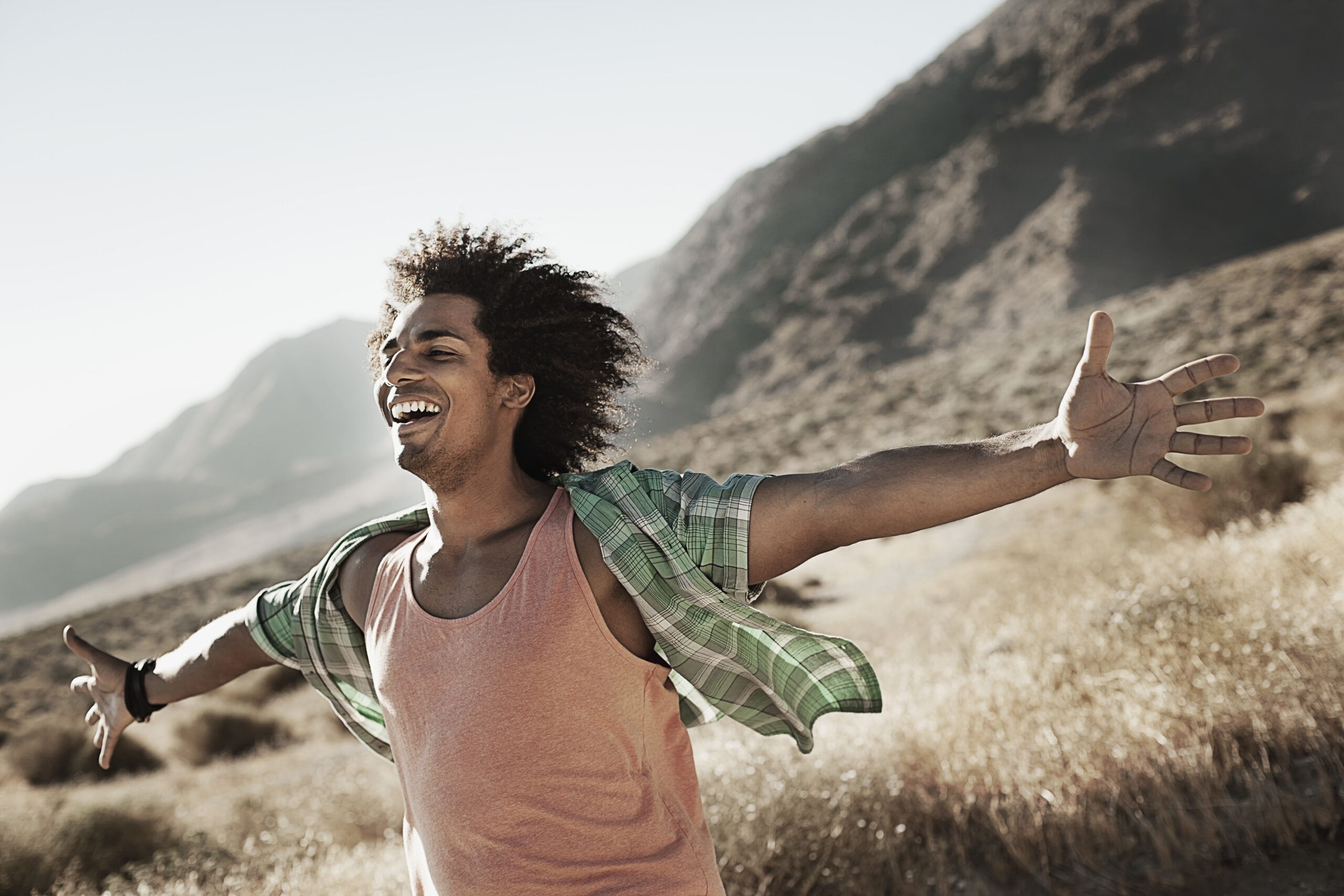 A man with light brown skin and dark curly hair smiles widely with his arms outstretched in front of a mountainous background.