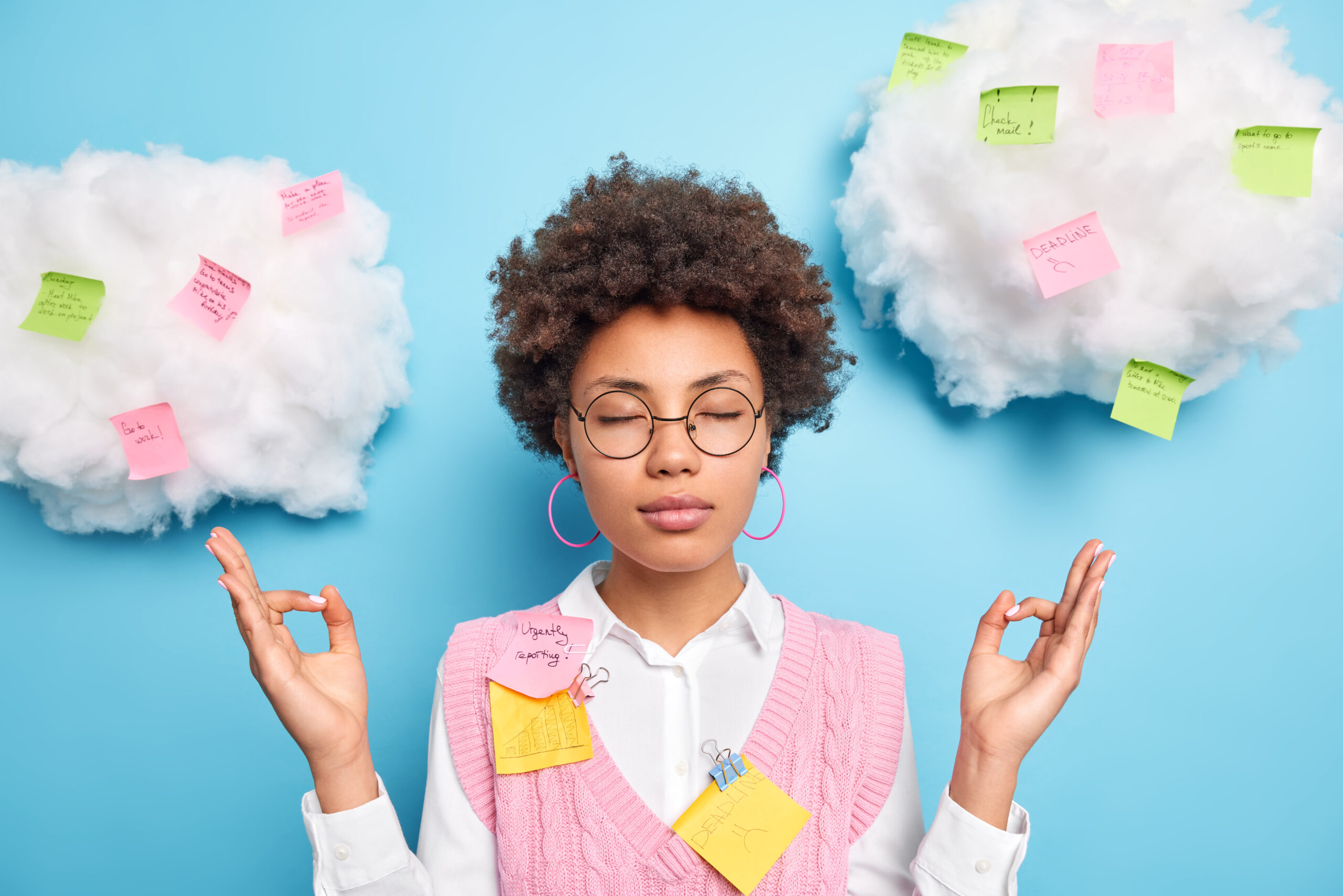 Brown skinned woman meditates with her eyes closed and her hands in a mudra position, in front of a blue background with two thought clouds that each have post-it notes with words and symbols on them.