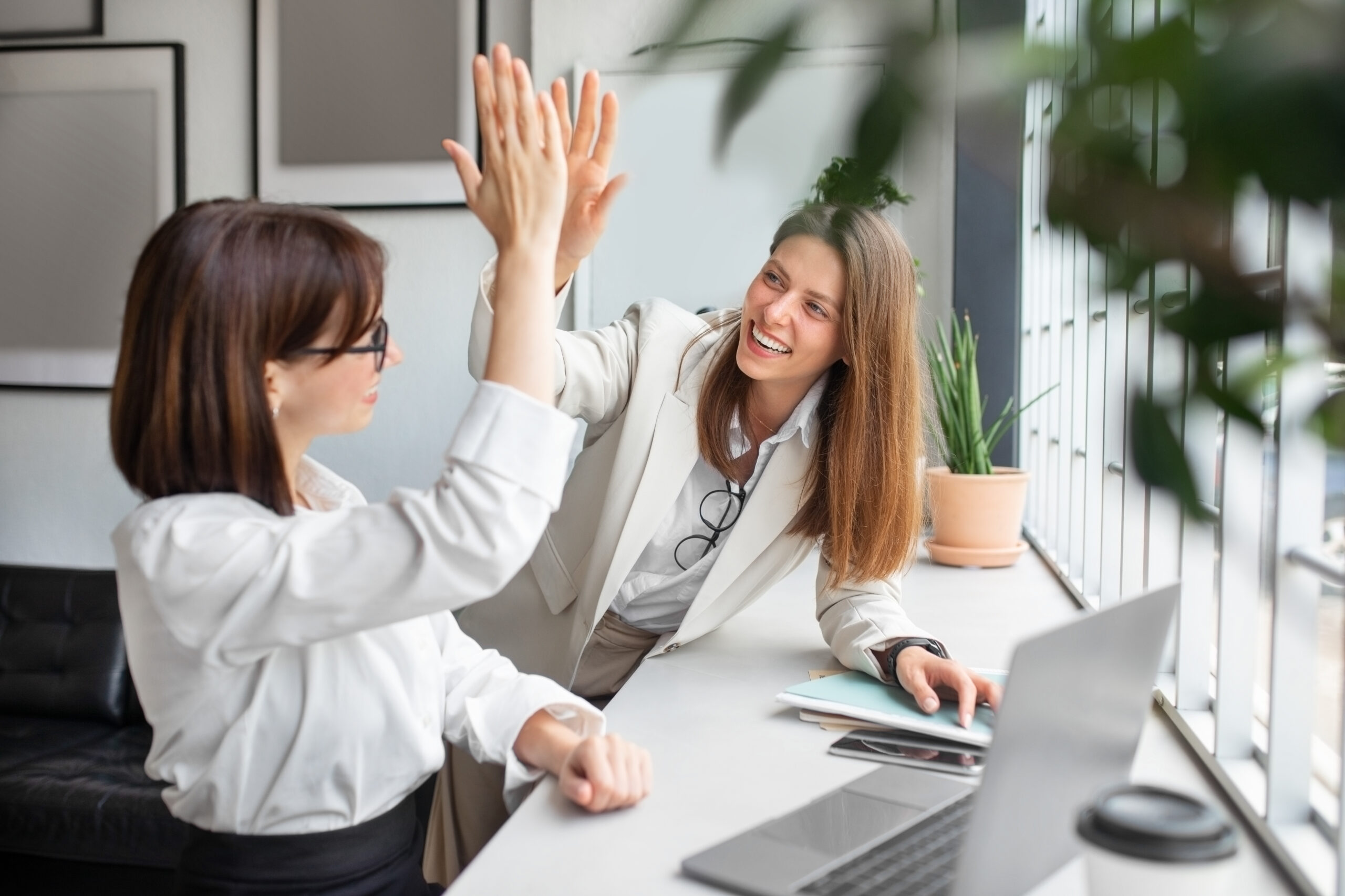 Photo of two white women in an office high-fiving with computers and office materials on the table in front of them.