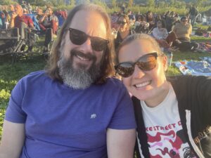 Image of Maegan, a white woman with dark hair and sunglasses, and Jonathan, a white man with long dark hair and beard and sunglasses, sitting on a lawn surrounded by other concert-goers.