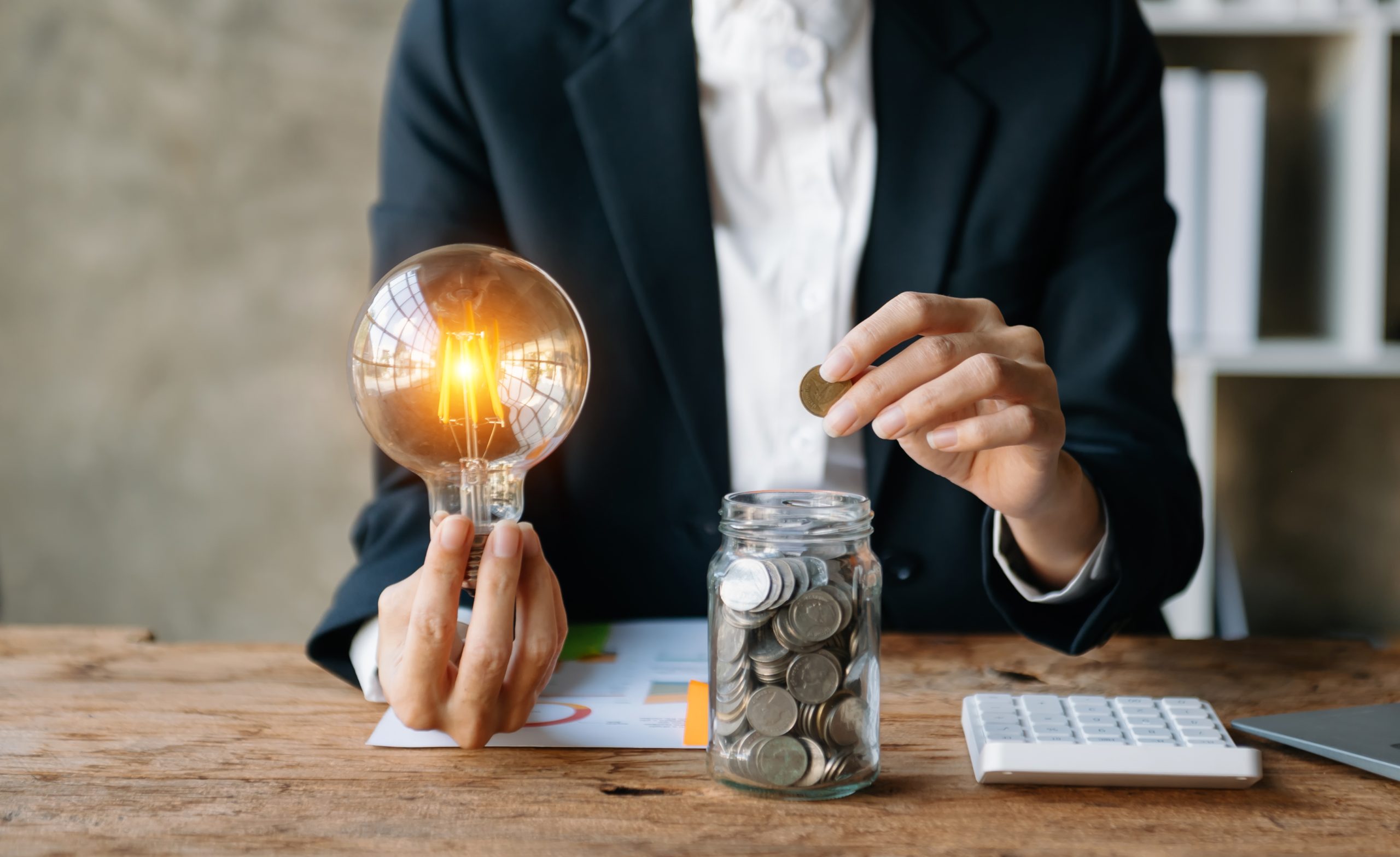 Image of a business woman with a lit lightbulb in one hand, putting coins into a jar with the other hand.