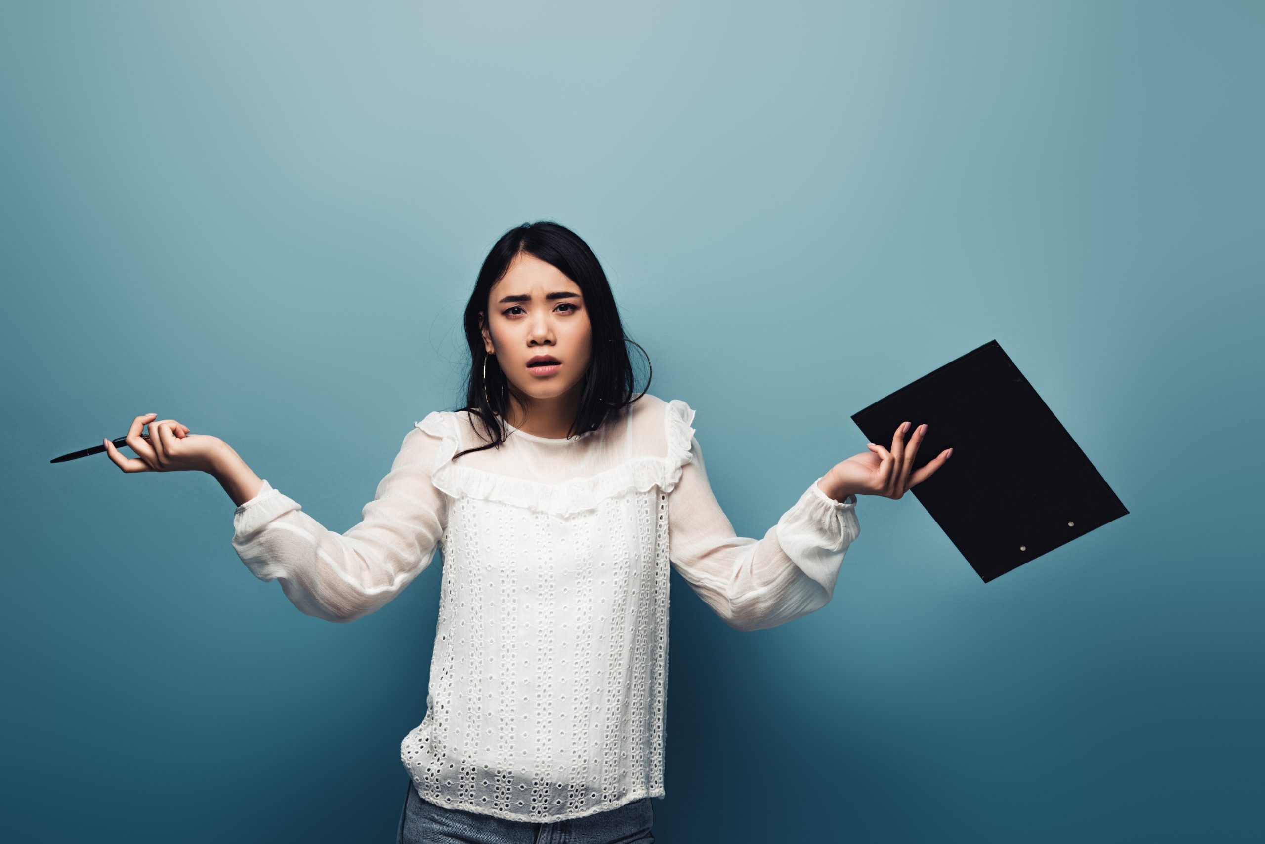 Asian woman with shoulder length hair and a white blouse, holding a pen in one hand and a clipboard in the other, arms in an "I don't know" gesture.