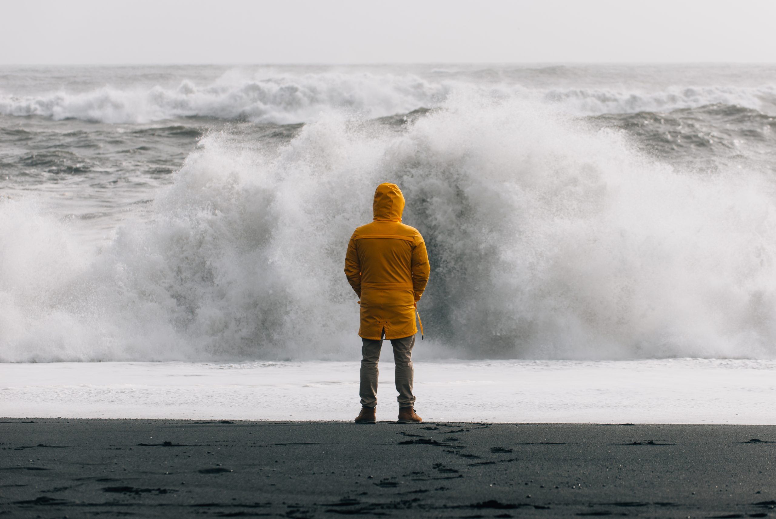 A solitary person in yellow rain coat standing with their back to the camera, facing crashing waves along the shore.
