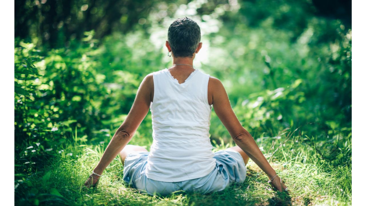 A figure with short, dark hair and a white tank top is seated in a cross-legged position on a patch of vibrant green grass. The individual faces away from the camera, their arms extended slightly to the sides, the blue bottoms barely visible. The tranquil nature setting is lush with green foliage, creating a serene and organic atmosphere.